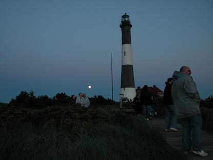 Moon rises behind darkened lighthouse, with group of people on boardwalk trail. Moon rises behind darkened lighthouse, with group of people on boardwalk trail.