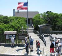 People on dock stroll toward bulletin board and small building, surrounded by lush green shrubs.