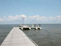 Long grey dock extends into calm bay water, beneath puffy white clouds in a clear blue sky.