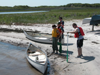 Canoeists prepare to board vessels on calm bay shoreline.
