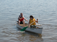Two canoeists paddle a small canoe on calm bay water.