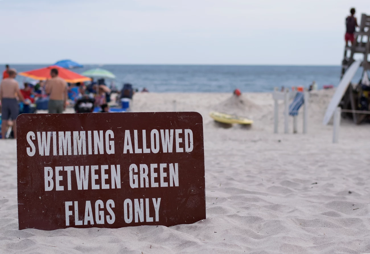 A sign reading, "Swimming allowed between green flags only" sits in the sand near a lifeguarded beach. A sign reading, "Swimming allowed between green flags only" sits in the sand near a lifeguarded beach.