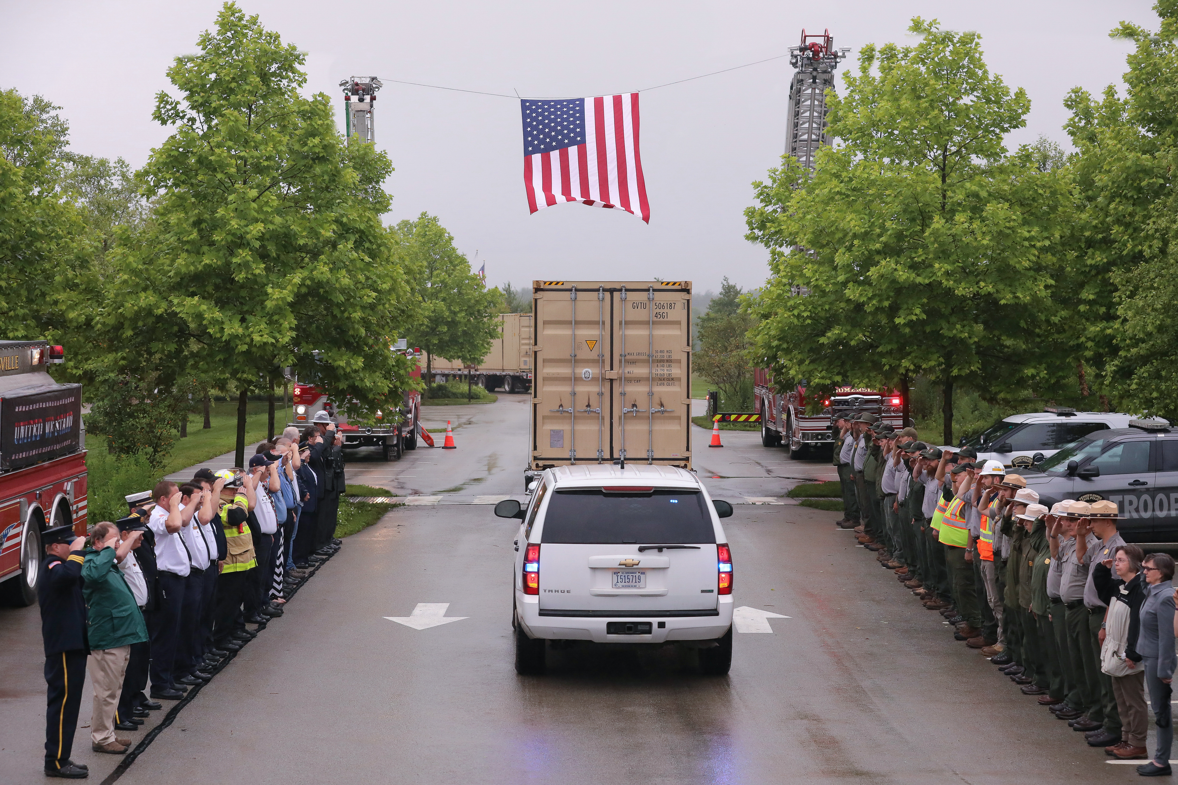 Flatbed truck with a large shipping container drives under a suspended American flag as onlookers salute.