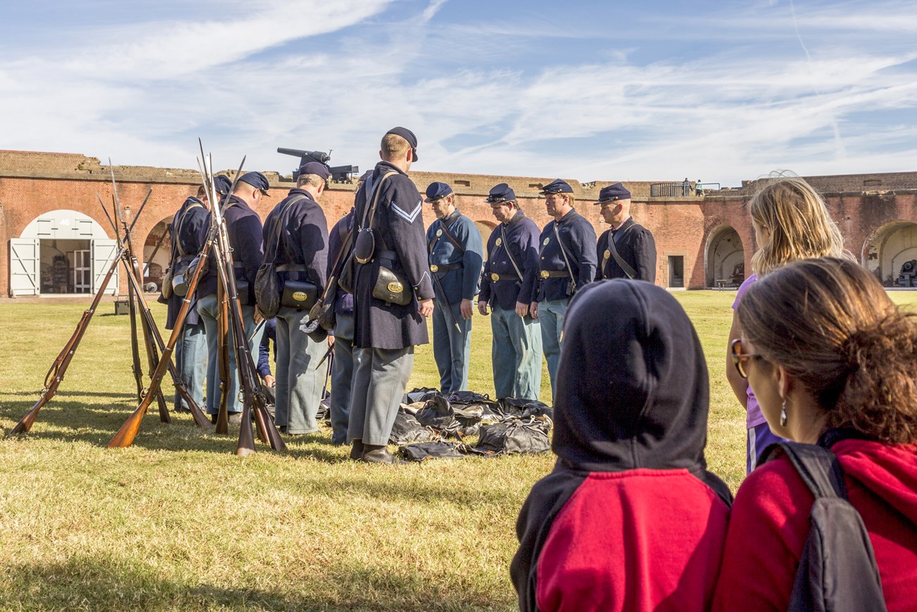 Visitors look on as a squad of 8 soldiers stand at attention while an officer inspects knapsacks.