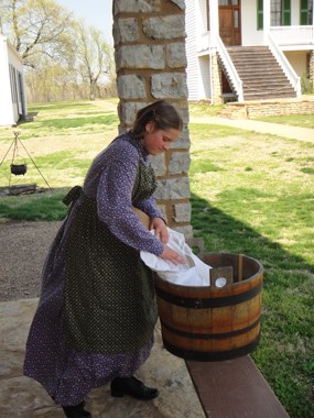 A laundress washes clothes in a basin under the awning of the barracks at Fort Scott
