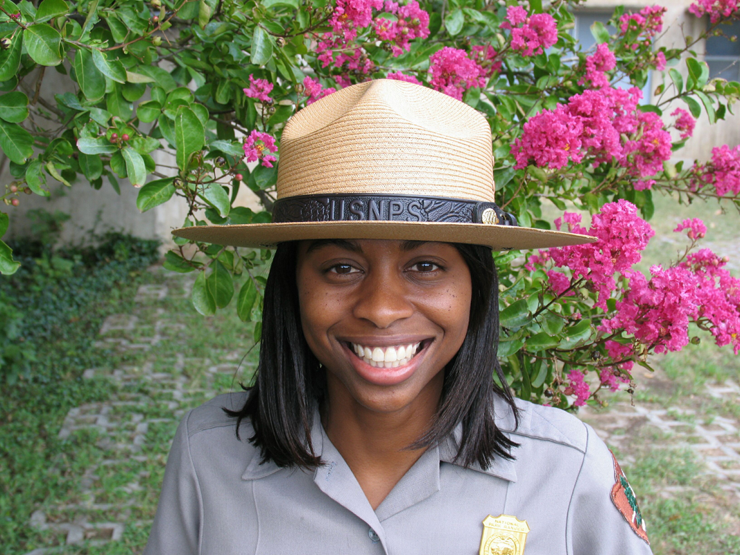 A smiling ranger wears a straw National Park Ranger hat. it has a wide brim and a brown leather strap wrapped around it.