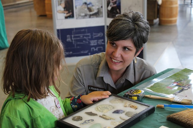 A park staff member kneels next to a little girl at a table covered in artifacts. She points explaining them to the girl.