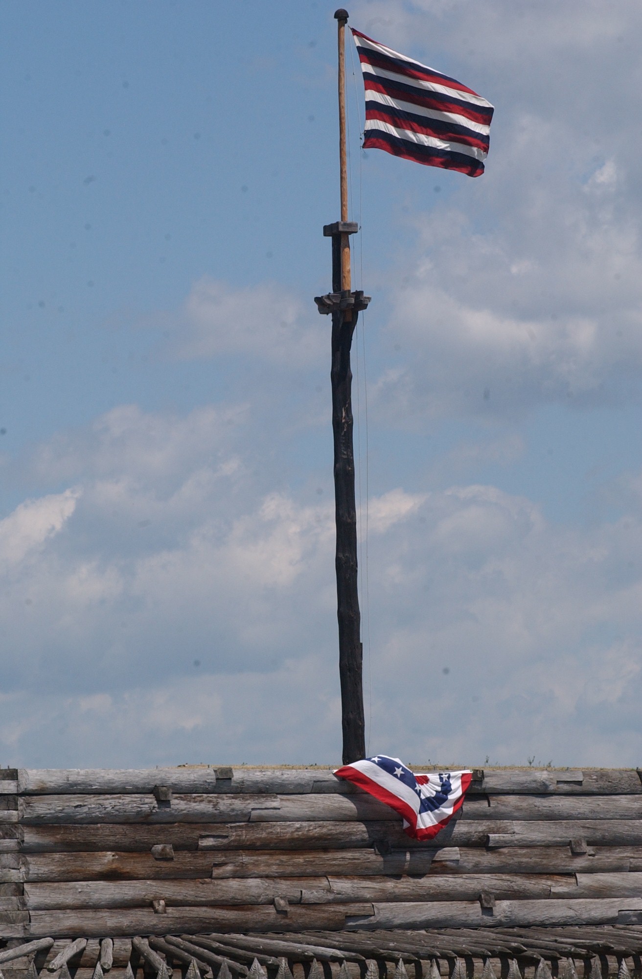 an alternating 13 stirped red, white, and blue striped flag flies in the sunlight over the fort wall