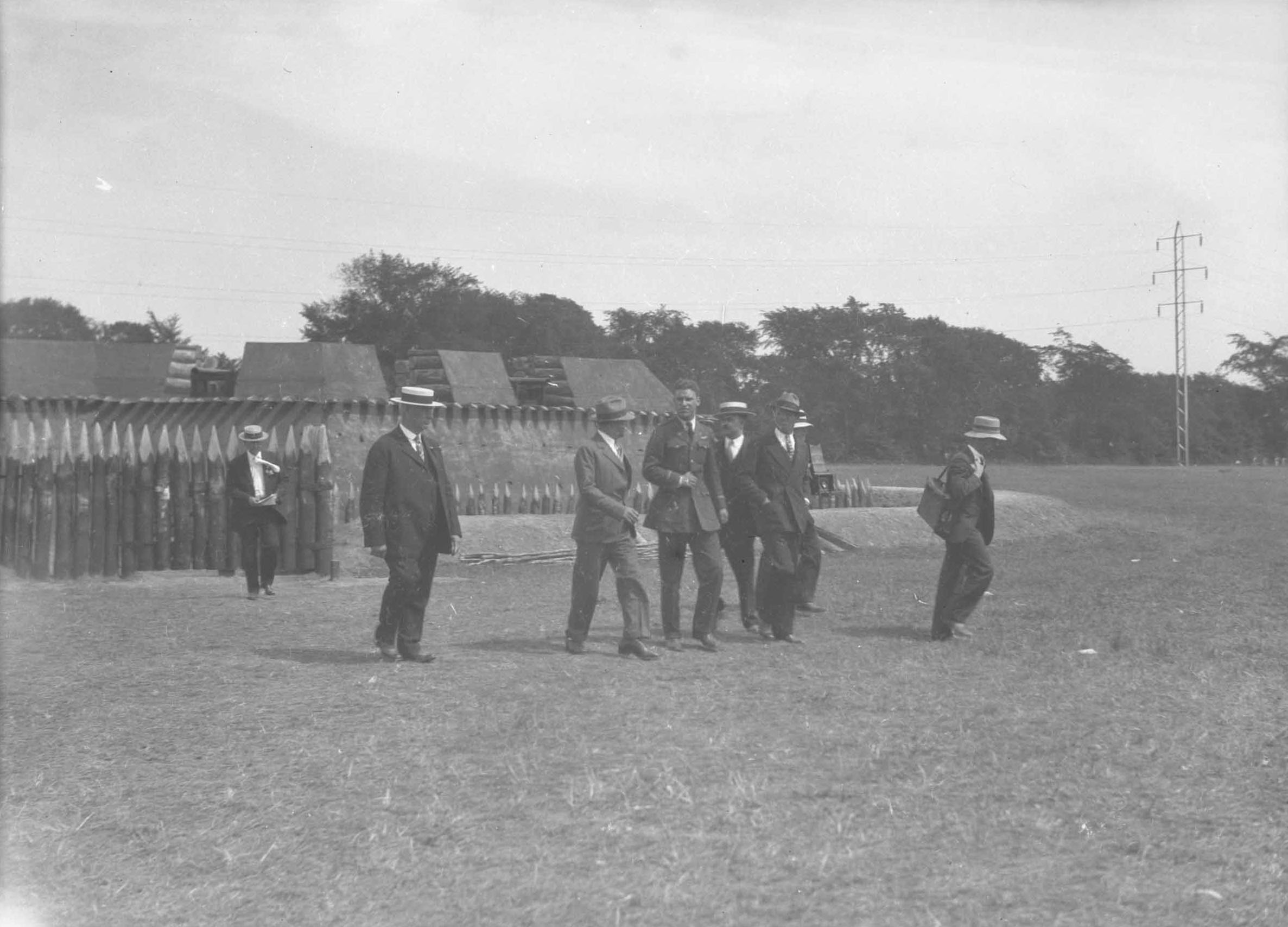 men in business suits walk in front of a tiny delicate looking fort with pickets and a ditch in the middle of a field