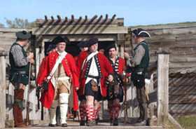 Under the shadow of the front gate, soldiers in kilts, white pants, and scarlet woolen jackets march towards a wooden bridge, framed by guards in green vests.