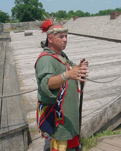 An Indian stands in the sunlight. He is holding a rifle. A blue beaded bag is slung over his shoulder. He is wearing a green baggy shirt and red tufts of feathers cover his bald head.