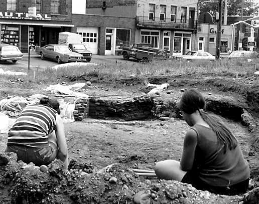 b&w, people sit in a trench, buildings surround them, you can see the remains of a foundation crumbling around them, they sort through it.