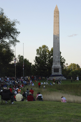 A white stone obelisk in shadow, people sit in a grassy green field surrounding, they are all shapes and colors