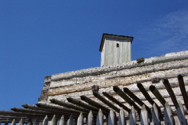 A stark greyish brown exterior of the fort in front, above you the cold blue sky. You are covered by the shadows of the pickets.