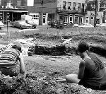 two women sit in a hole digging near bricks, a cityscape behind them