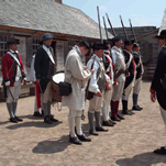 men in white shirts and blue and red wool coats stand in a line in a dusty open area