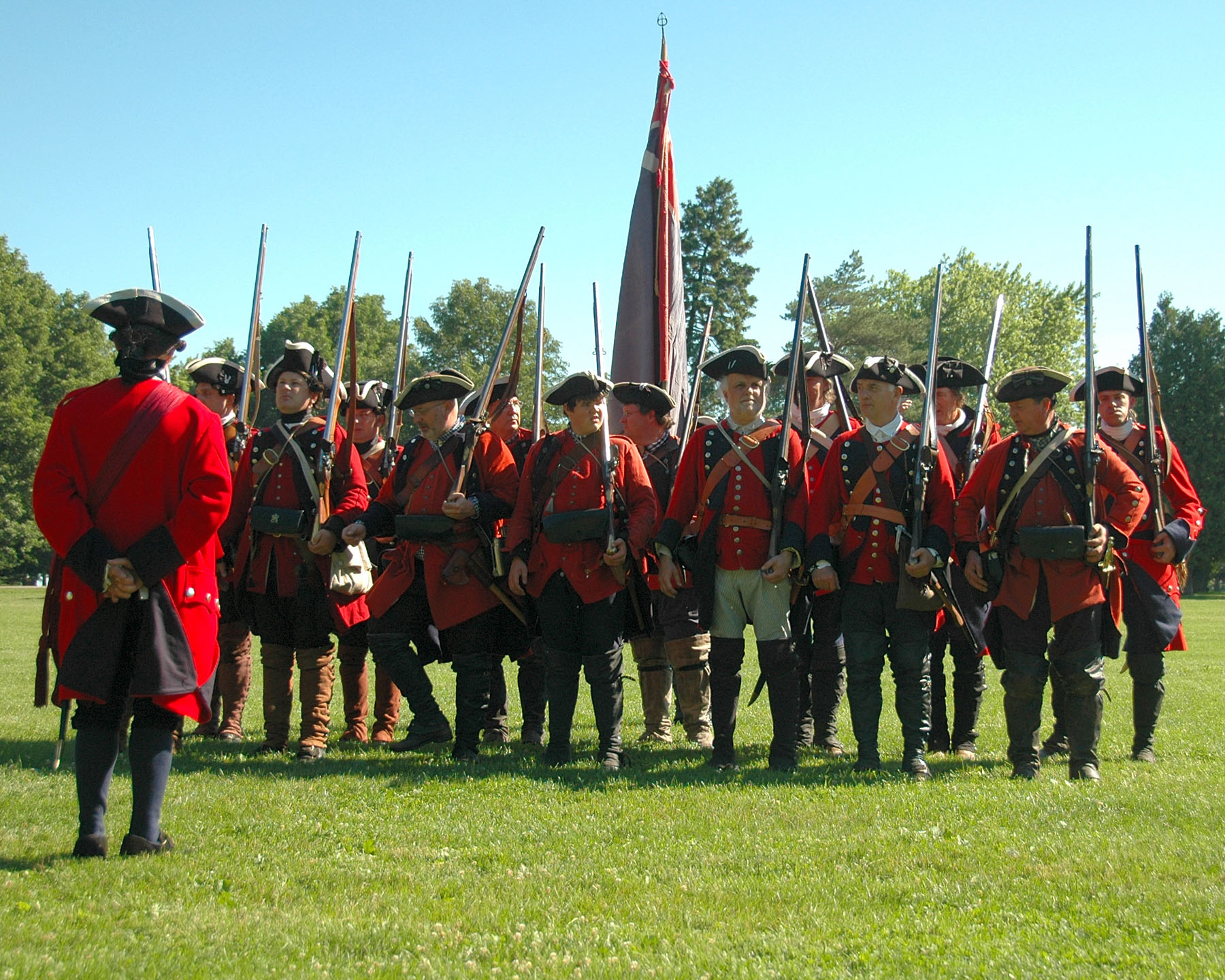 men in red jackets stand in a line with muskets.