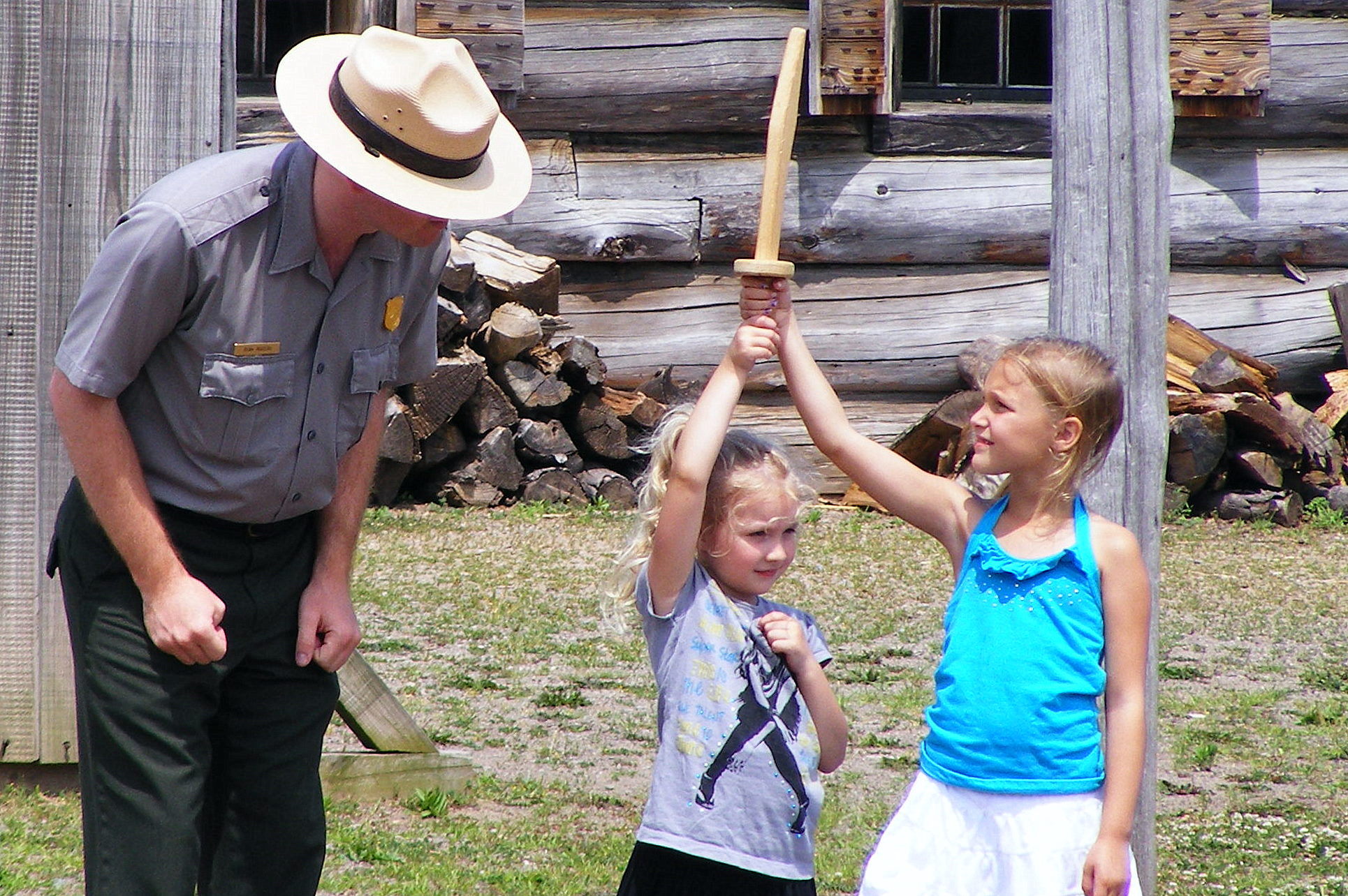 A little girl and her sister hold a wooden sword next to a park ranger.