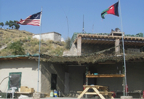 a historic red, white, and blue flag flies on a pole near a dusty tan mountain and a concrete building; next to it a striped green, red, blue flag