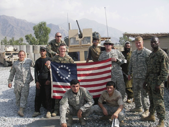 U.S. Army men is camouflage stand in a grey/tan rocky colored area surrounding a flag.blue flag.