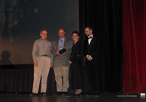 four people on stage, they smile widely, all hold a piece of a cirular object, their images projected on a screen behind.