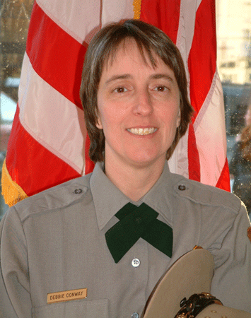 a woman with short brown hair smiles widely. she wears a grey uniform, green tie and stands in front of an American flag.