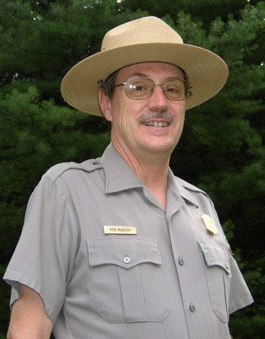 a man in a straw park ranger hat looks up and smiles in front of green trees