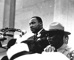 A man in a suit stands stern in front of a microphone in between great pillars