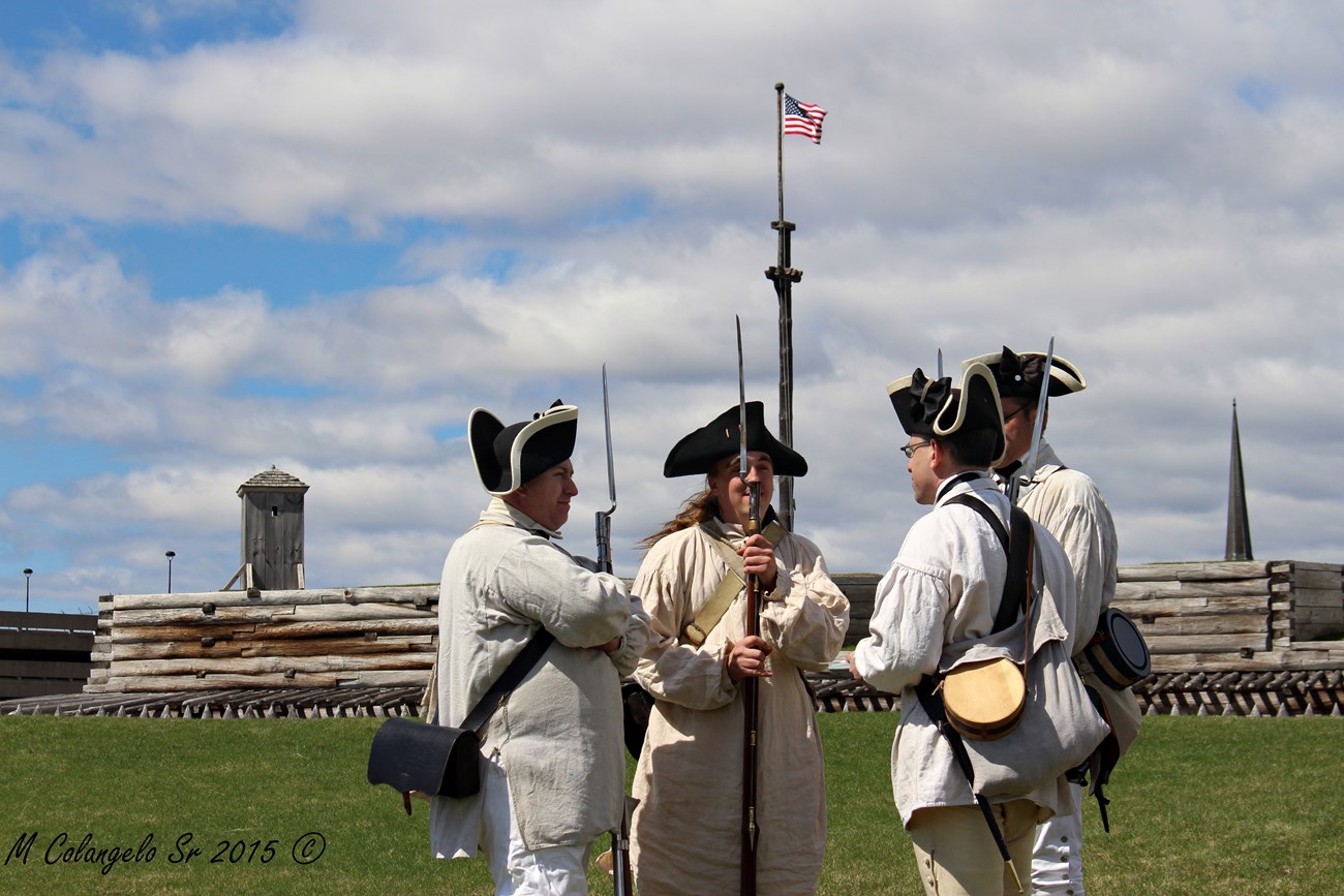 Continental soldiers stand in a circle with muskets at their sides. The fort sits in the background.