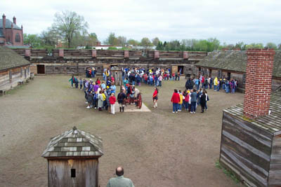 A view from a building inside the fort overlooks the vast dirt covered parade ground. On it, various people in multi-colored clothing stand in a square participating in a program, all talking to each other. They seem very tiny from this distance, and a chimney blocks part of the view.