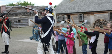 in a neat row children in colorful jackets mimic the motions of a soldier in the background