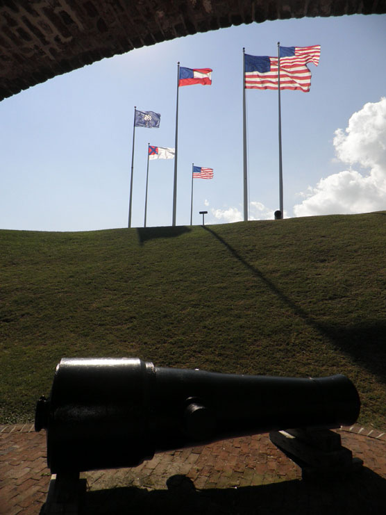 A Civil War cannon with the U.S. flag and five historic flags of Fort Sumter flying in the distance.