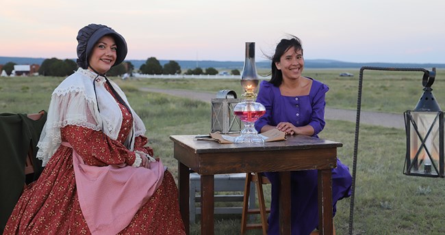 two women dressed in 19th century dresses sitting in front of oil lamp