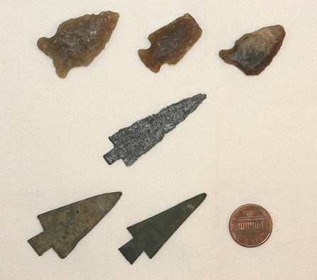 Three sharpened stone triangles and three rusty sharp tipped metal triangles with a penny that is about half the size of each triangle.