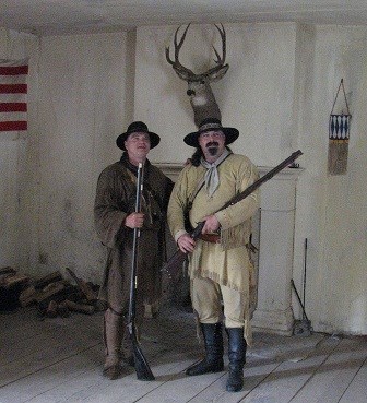 Two men wearing historic leather clothes holding rifles in front of a fireplace with a buck head mounted above them.