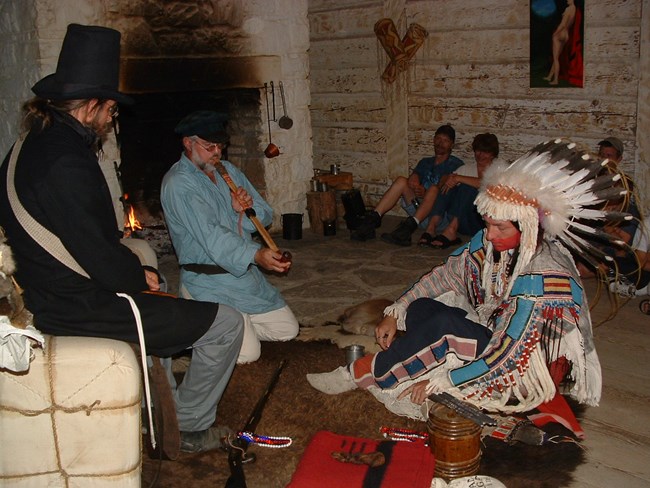 A man in traditional American Indian Dress sitting on a brown buffalo robe. A man in historic dress is kneeling next to him smoking a long pipe. Another man wearing dark historic dress and a black hat sits on a tan bale beside them.