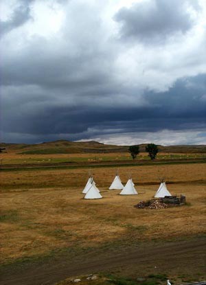 Five tipis in a green and brown grassy field with very dark clouds overhead.