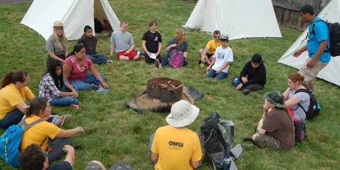 Campers sit on a circle in the grass outside with Park Rangers and OMSI instructors, with tents in the background.