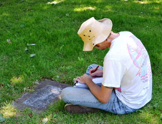 Student sitting and recording information in a notebook from a grave marker.
