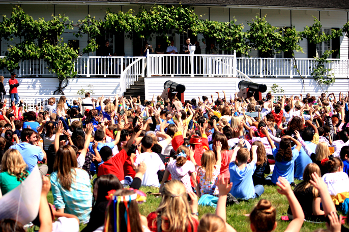 Image of third grade students sitting on the lawn in front of the Chief factor's House, listening to the superintendent talk about the significance of the site. Photo is from the perspective of students sitting, and shows several hundred of them raising their hands in response to a question from the speaker, located near the center of the image.