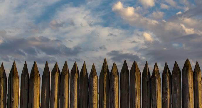 Image of the pointed tops of the stockade wall, with a background of bright blue sky and clouds.