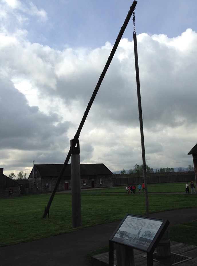 Photo of the well sweep at Fort Vancouver, with a wayside panel in front and a building in the background.