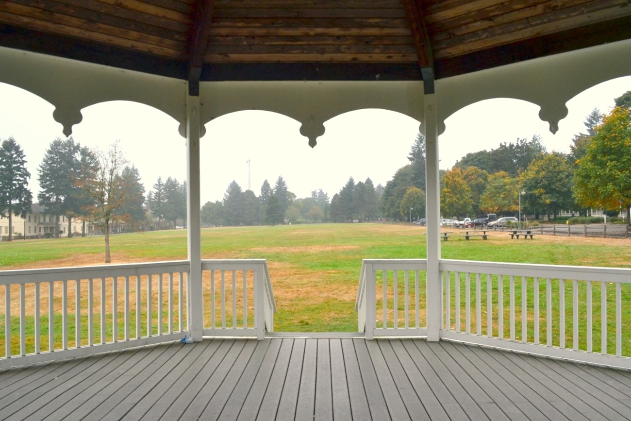 The view from inside the Bandstand. The photographer is standing inside a round, covered wooden bandstand, facing the bandstand entrance, which has a short flight of stairs down to a grassy field.
