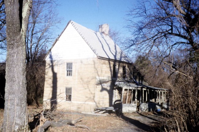 A two and a half story home seen from the side. The house is built of wood and covered in stone. It has a steeply pitched roof and a dilapidated front porch. It stands in a forested area.