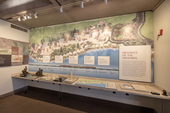 A large watercolor drawing of an ariel view of the monument is displayed on the wall. A timeline of FDR'S terms as President is displayed near the bottom of the painting. Below the timeline is a table with various tactile, audio, & braille exhibits.