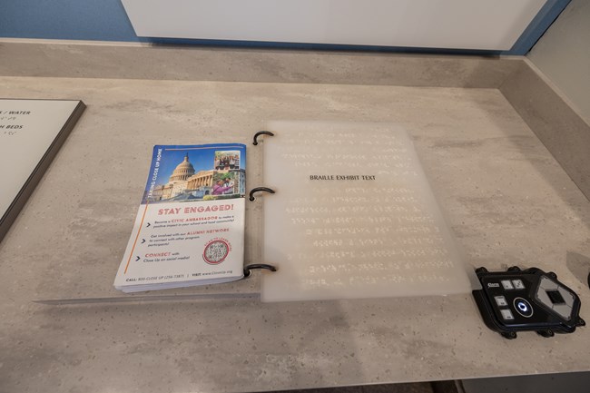 A clear booklet with braille is attached to a three ringed binder. To the right is an audio option to listen to the exhibit.