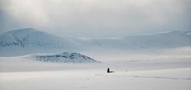 A musher in a distant and vast snowy tundra landscape with mountains and sunlight shining down into the valley in the distance