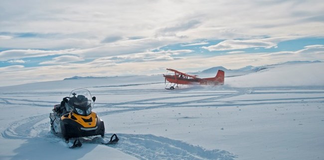 A ski-equipped bushplane lands on the snowy arctic tundra next to a snowmachine