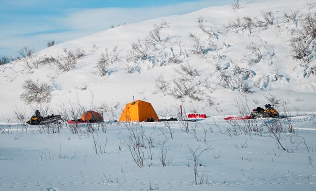 A snowy winter camp with a bright yellow tent and two snowmachines