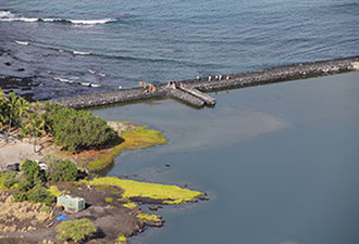 People stand on a rock wall that separates the ocean from the calm waters of the Kaloko Fishpond